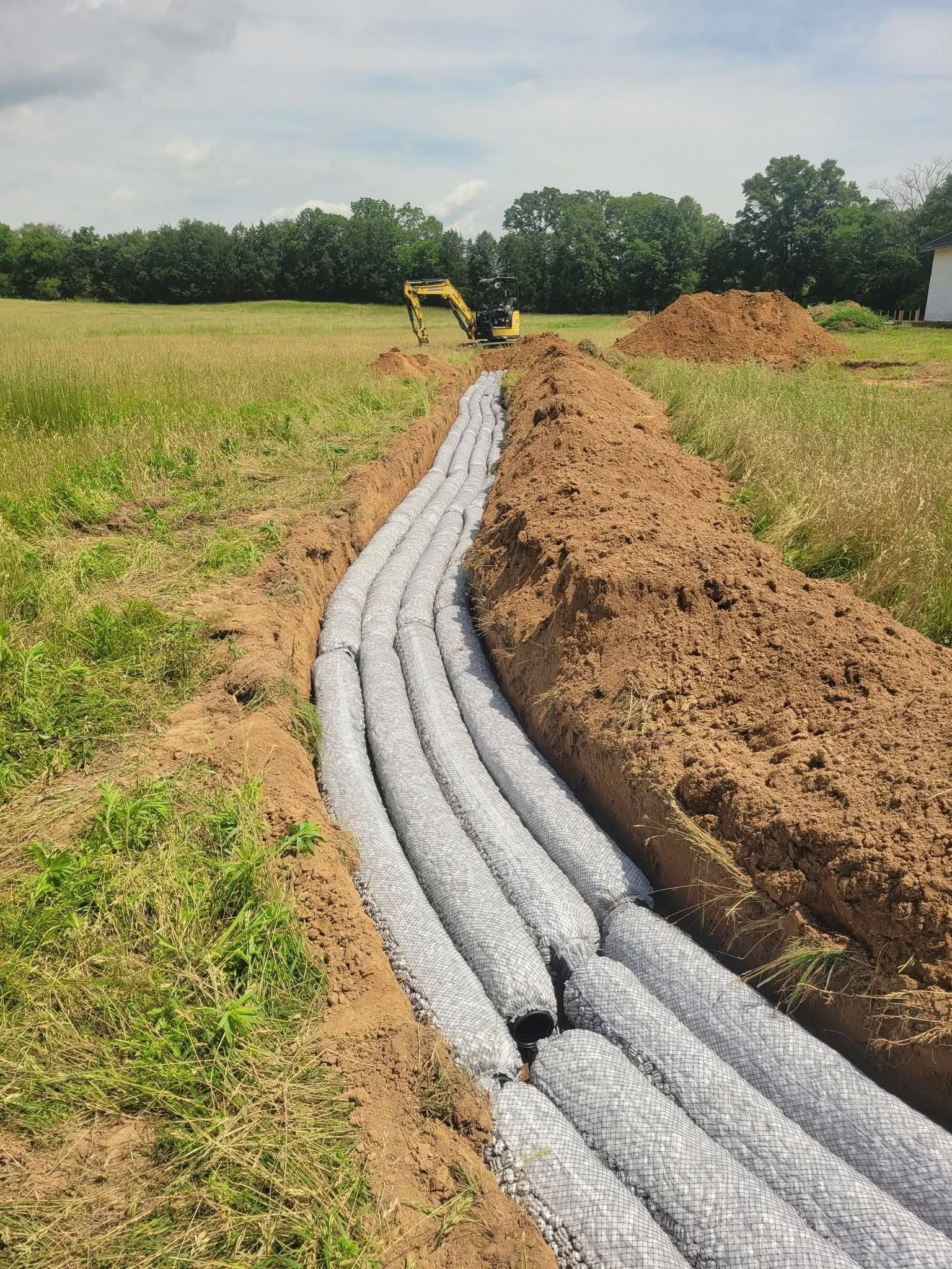 Sandbags line a trench in a grassy field, with an excavator and dirt piles in the background.