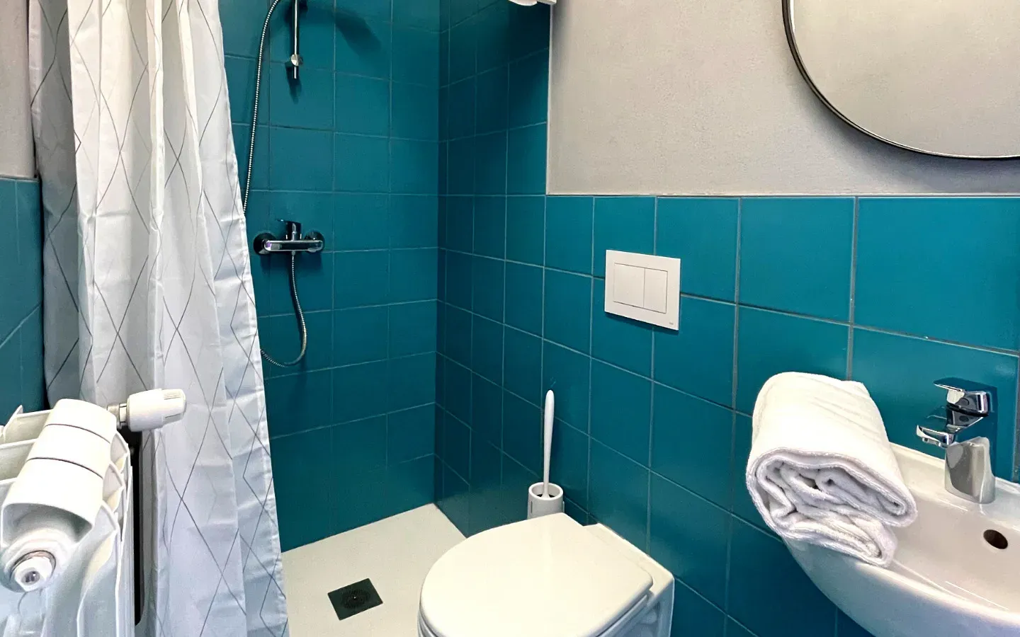 Bagno piastrellato color verde acqua con doccia, WC, lavandino, specchio. Asciugamano bianco.