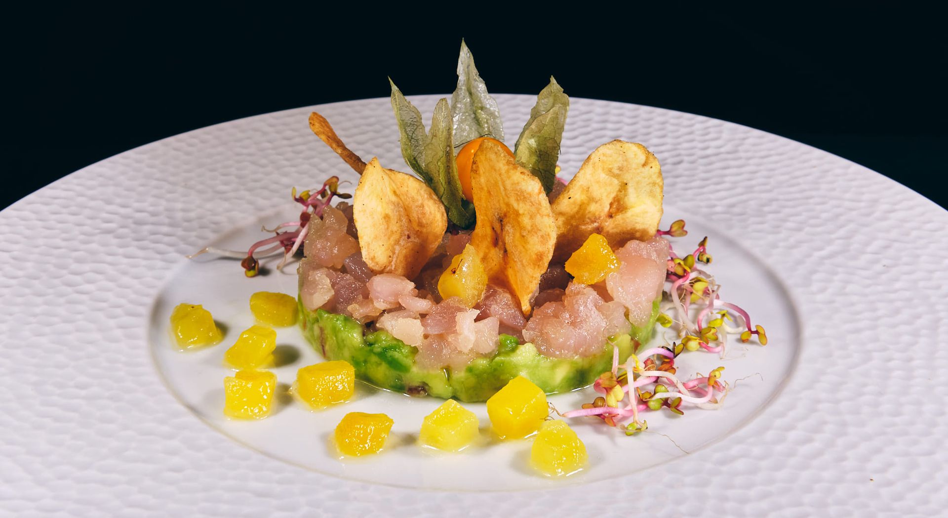 Tartare di frutti di mare su avocado, guarnita con spicchi d'arancia, patatine e germogli su un piatto bianco.