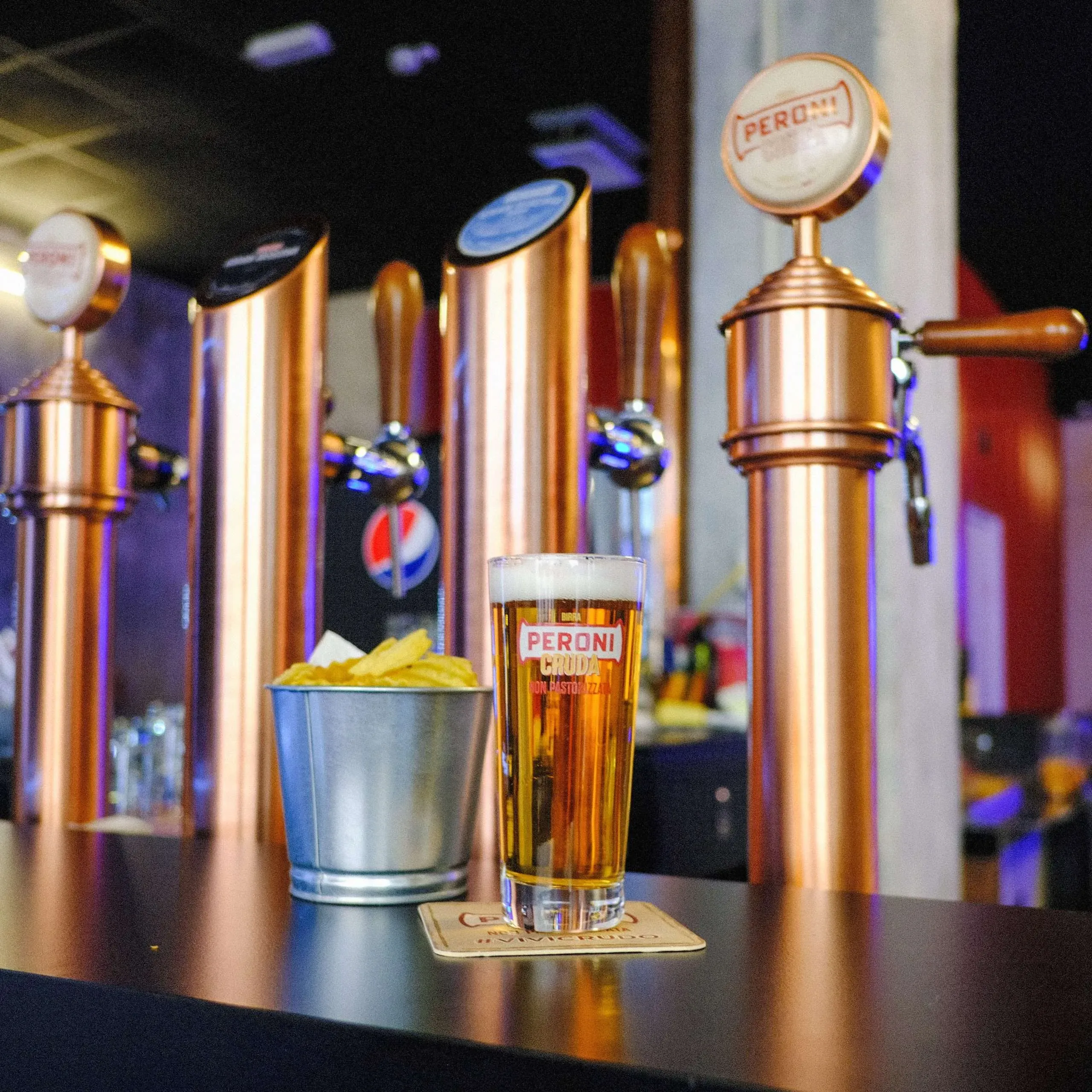 Beer glass and snacks on a bar, with copper-colored beer taps in the background.