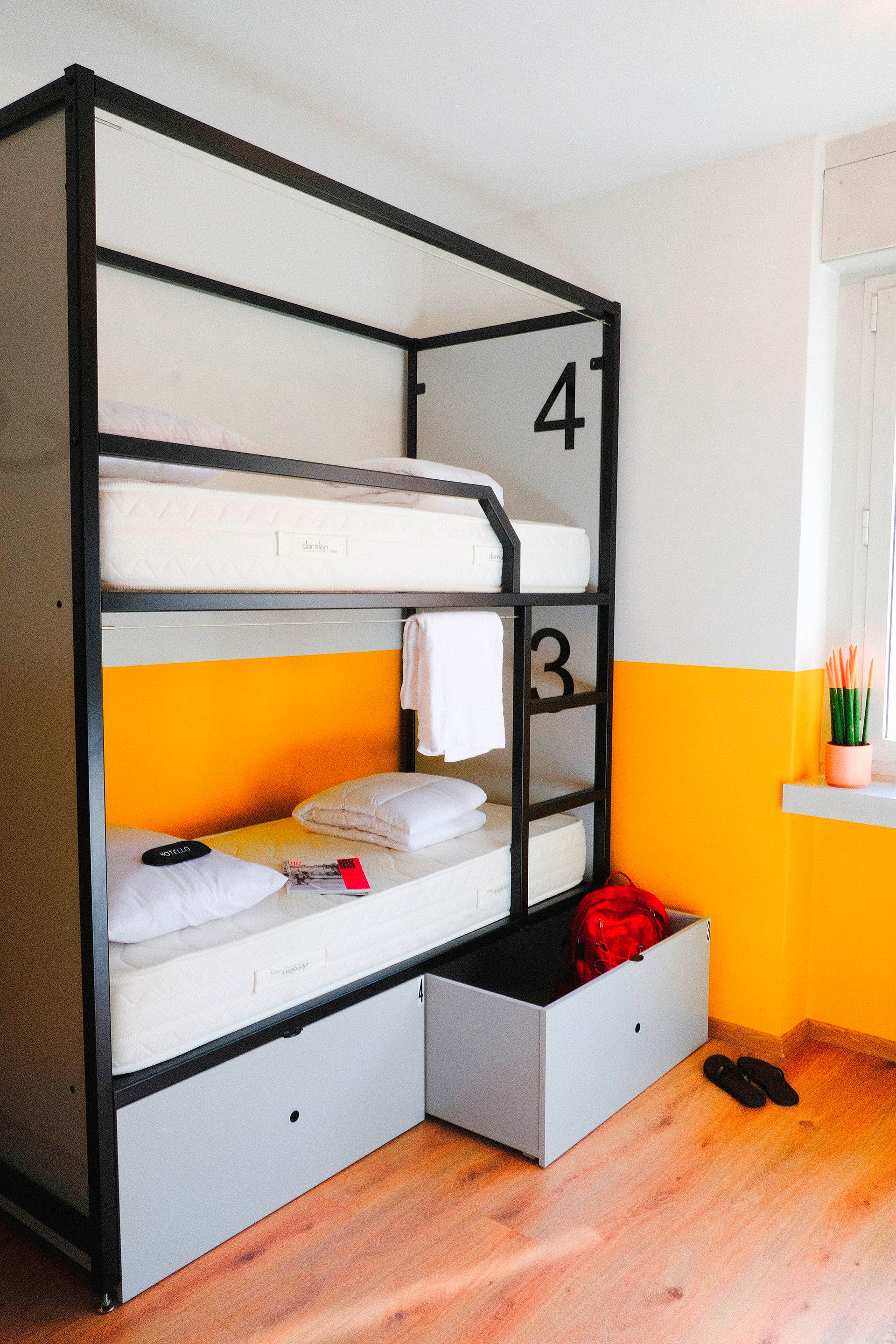 Letto a castello in una stanza d'ostello. Pareti arancioni e bianche, struttura nera, cassetti grigi.