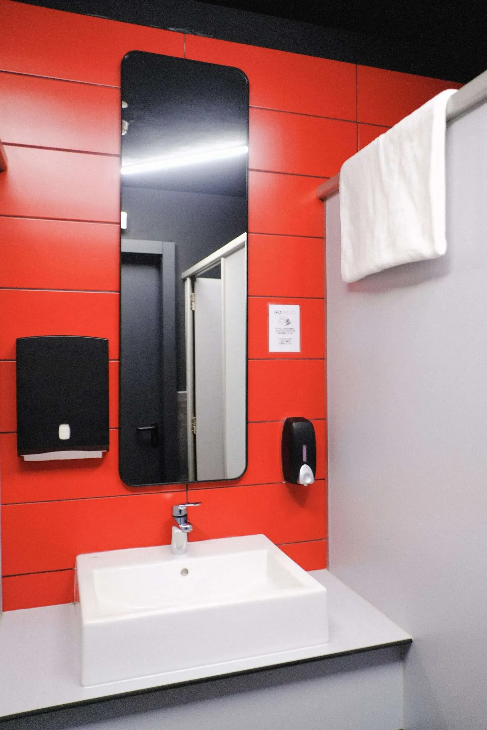 Bagno rosso e grigio con lavandino, specchio, distributore di carta assorbente e asciugamano su barra.