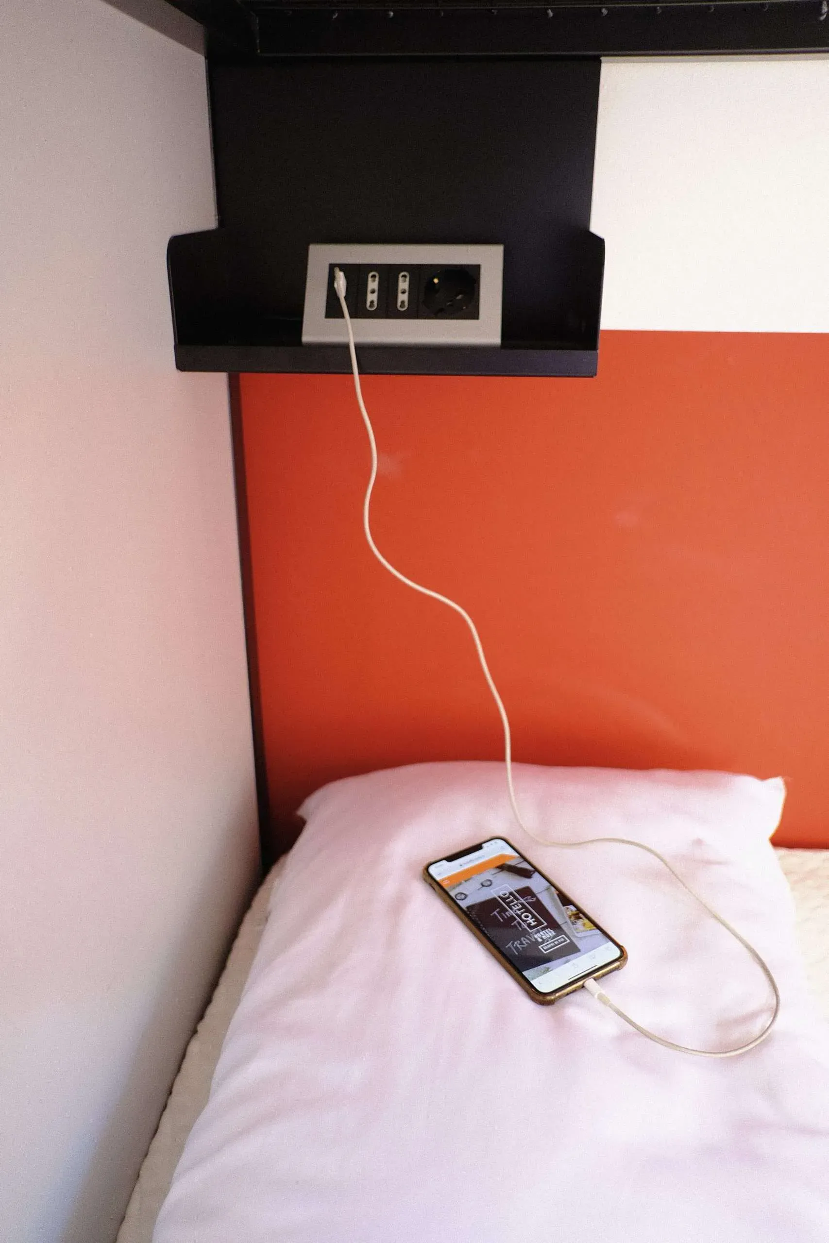 Un telefono in carica su un cuscino; presa elettrica in alto. Cavo bianco, parete arancione, mensola nera.