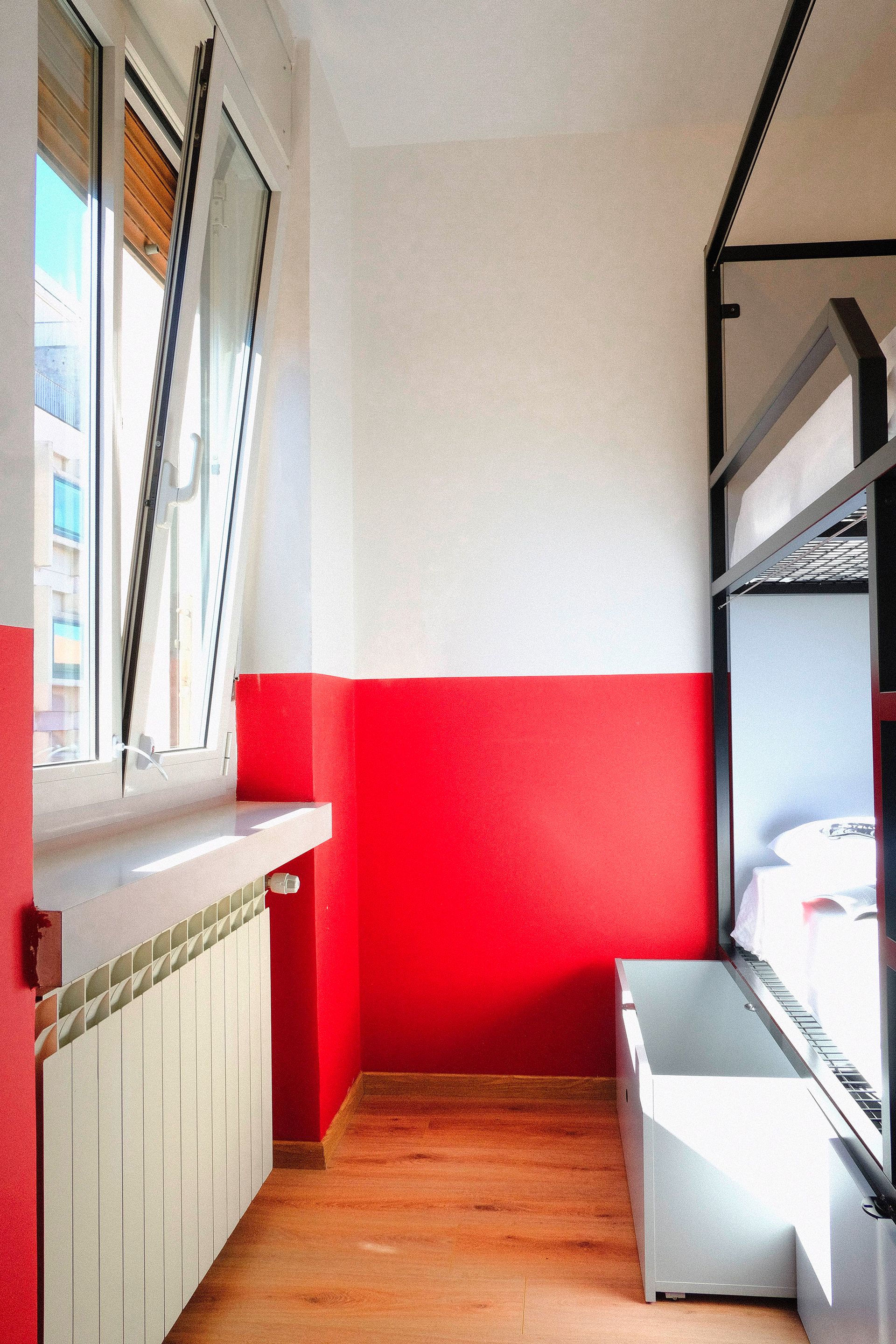 Camera rossa e bianca in stile ostello con letto a castello, finestra, termosifone e pavimento in legno.