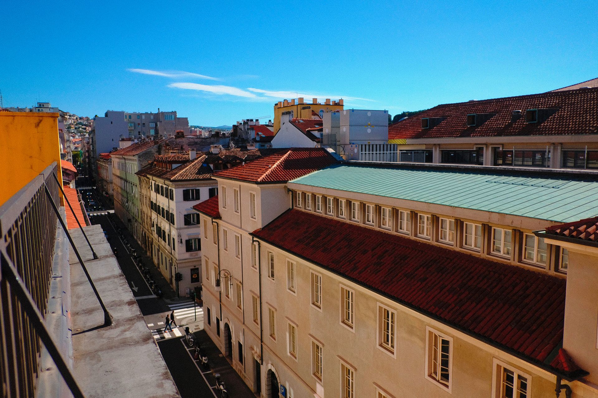 Veduta della città con edifici, tetti di tegole rosse, cielo azzurro soleggiato e qualche persona che cammina.