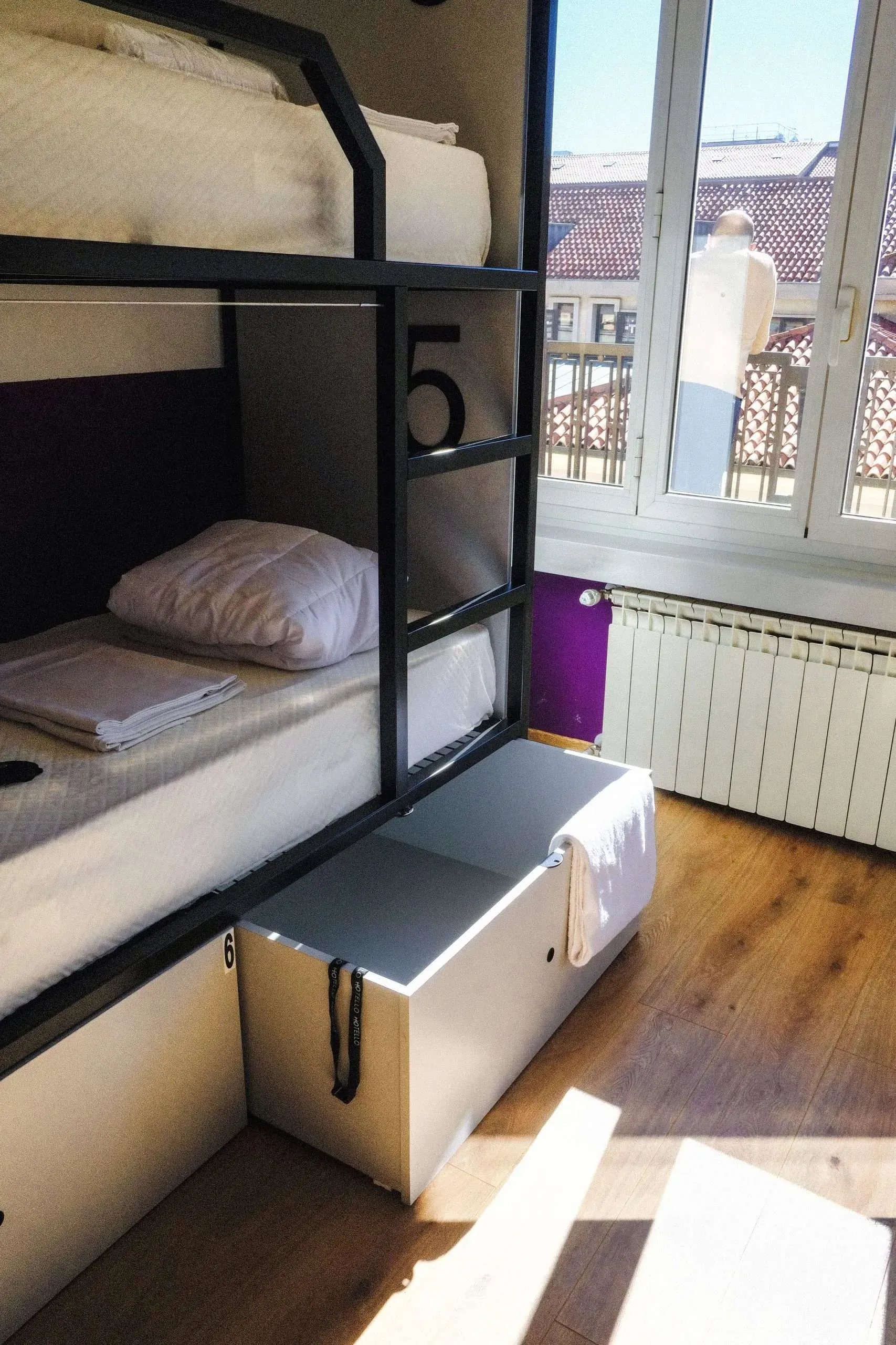 Letti a castello in una stanza di un ostello con finestra, pavimento in legno e un baule bianco.