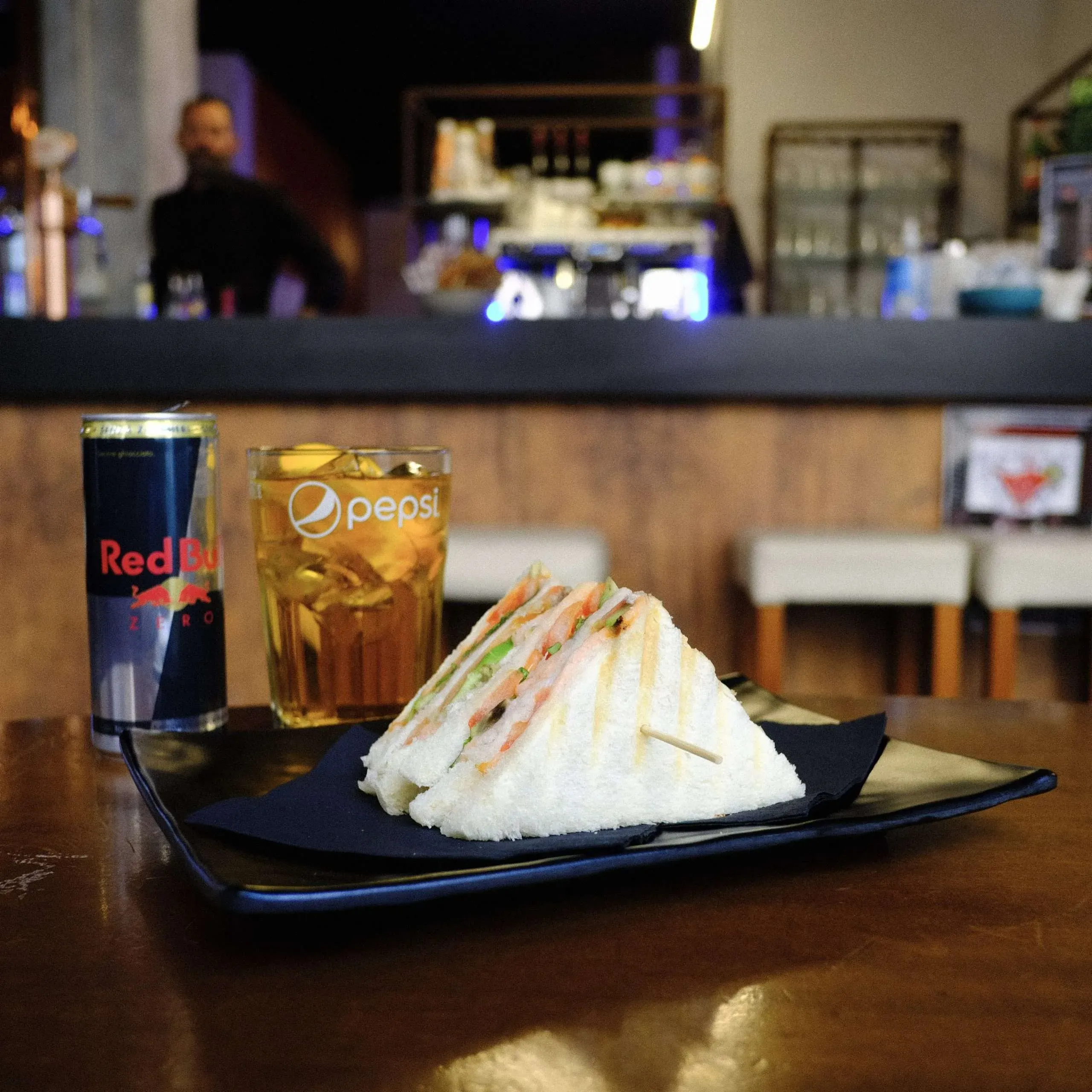 A sandwich, Pepsi, and Red Bull on a table in a bar, with a bartender in the background.