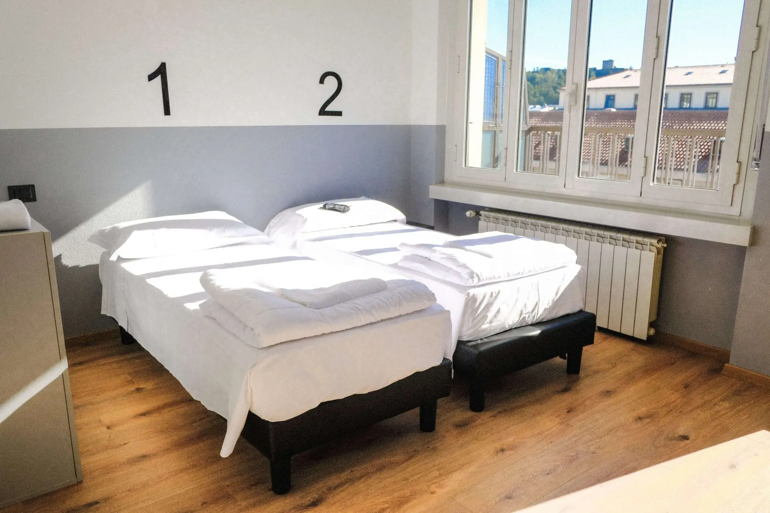 Camera da letto con due letti singoli, biancheria bianca, ampia finestra, numeri 1 e 2 sulla parete.