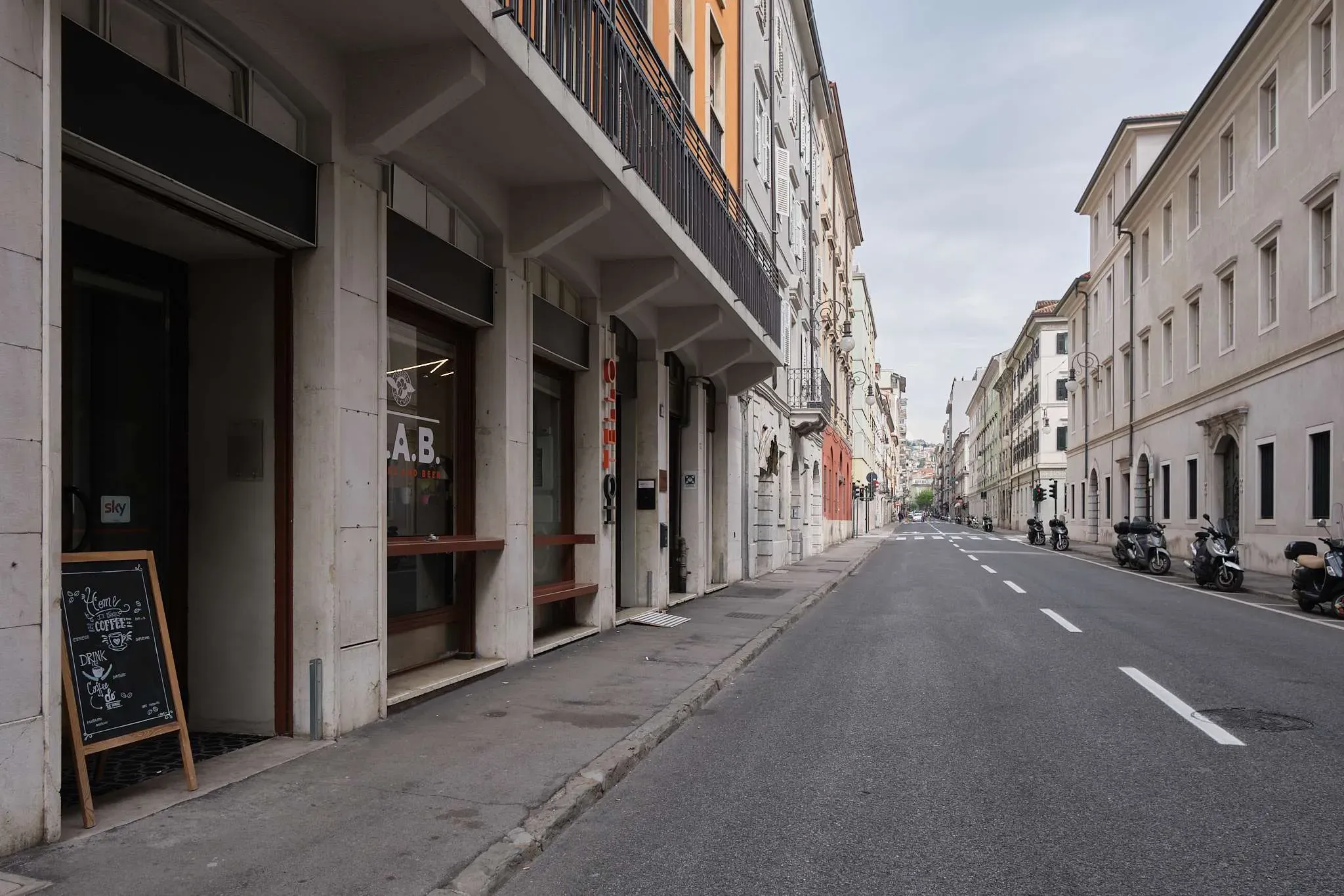 Strada cittadina deserta con vecchi edifici, un cartello con la lavagna e motociclette parcheggiate.