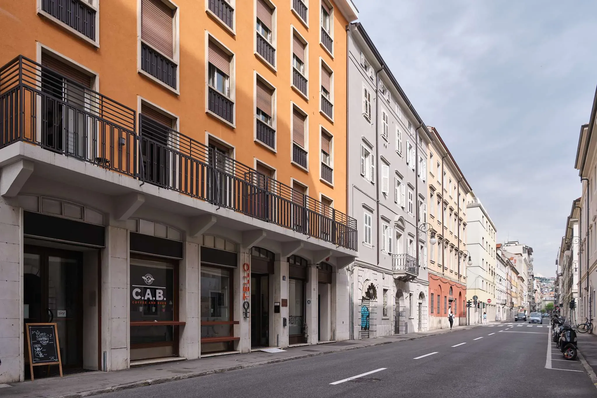 Scena di strada con edifici colorati, alcuni con balconi. Un bar al piano terra. Strada deserta.