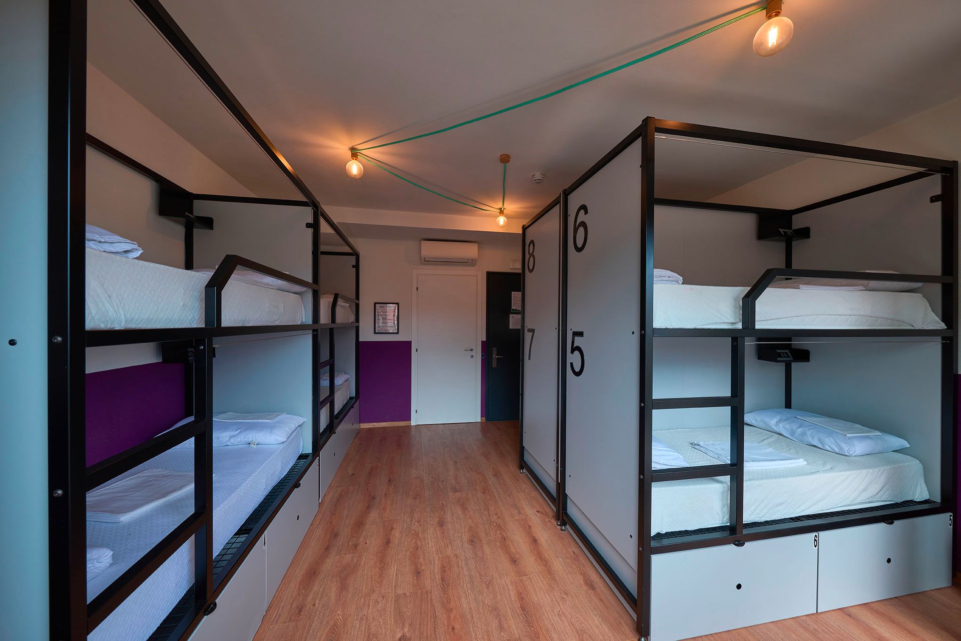 Camerata in ostello con letti a castello, numeri sui divisori, pavimenti in legno, pareti viola e bianche, luci sospese.