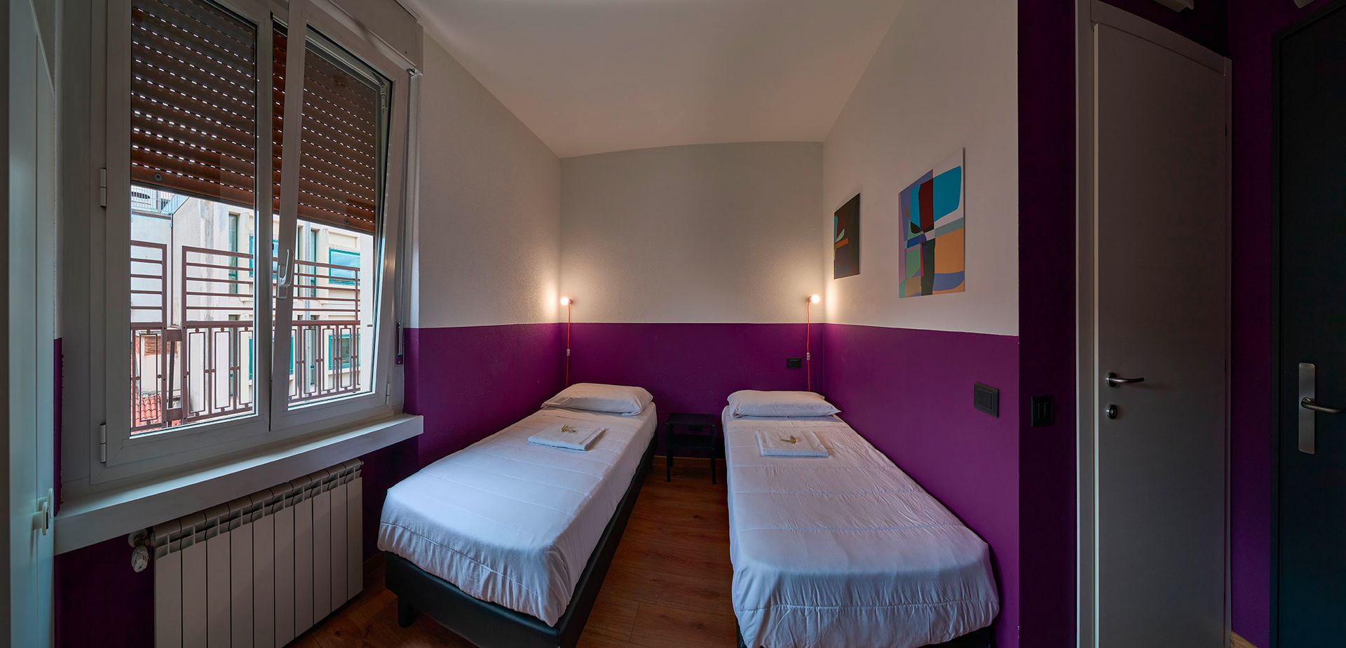 Una piccola stanza d'albergo con due letti singoli, dipinta di viola e bianco. Una finestra a sinistra. Una porta a destra.