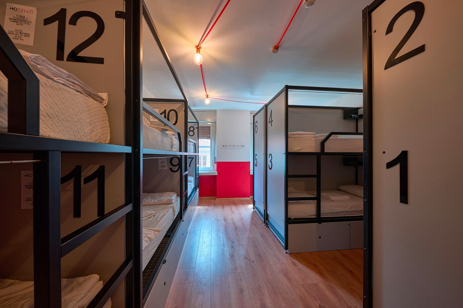 Camerata con letti a castello numerati, dettagli rossi e uno stretto passaggio.