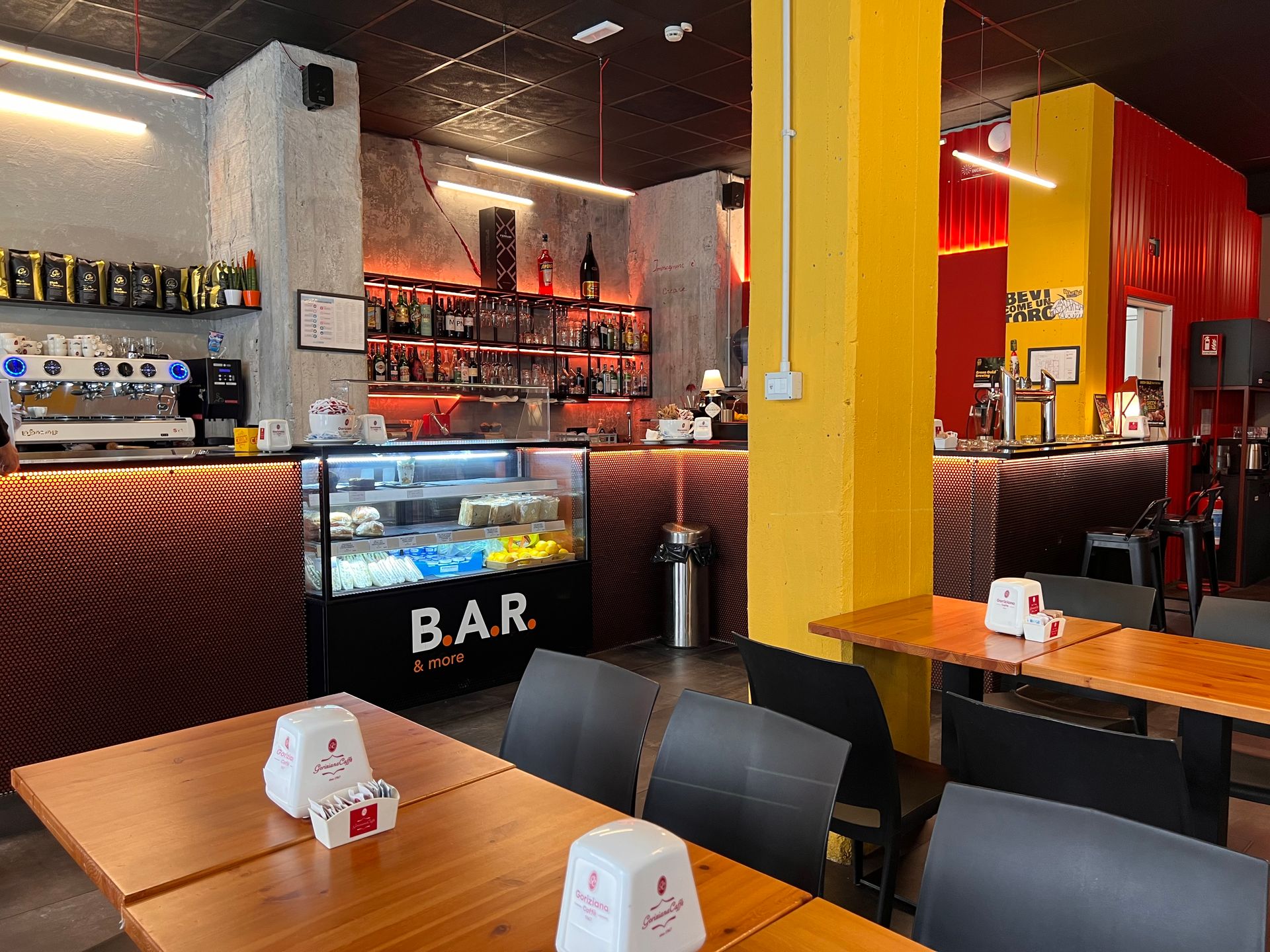 Vista interna di un bar con tavoli, bancone e scaffali pieni di bevande. Pareti gialle e rosse, tavoli in legno.