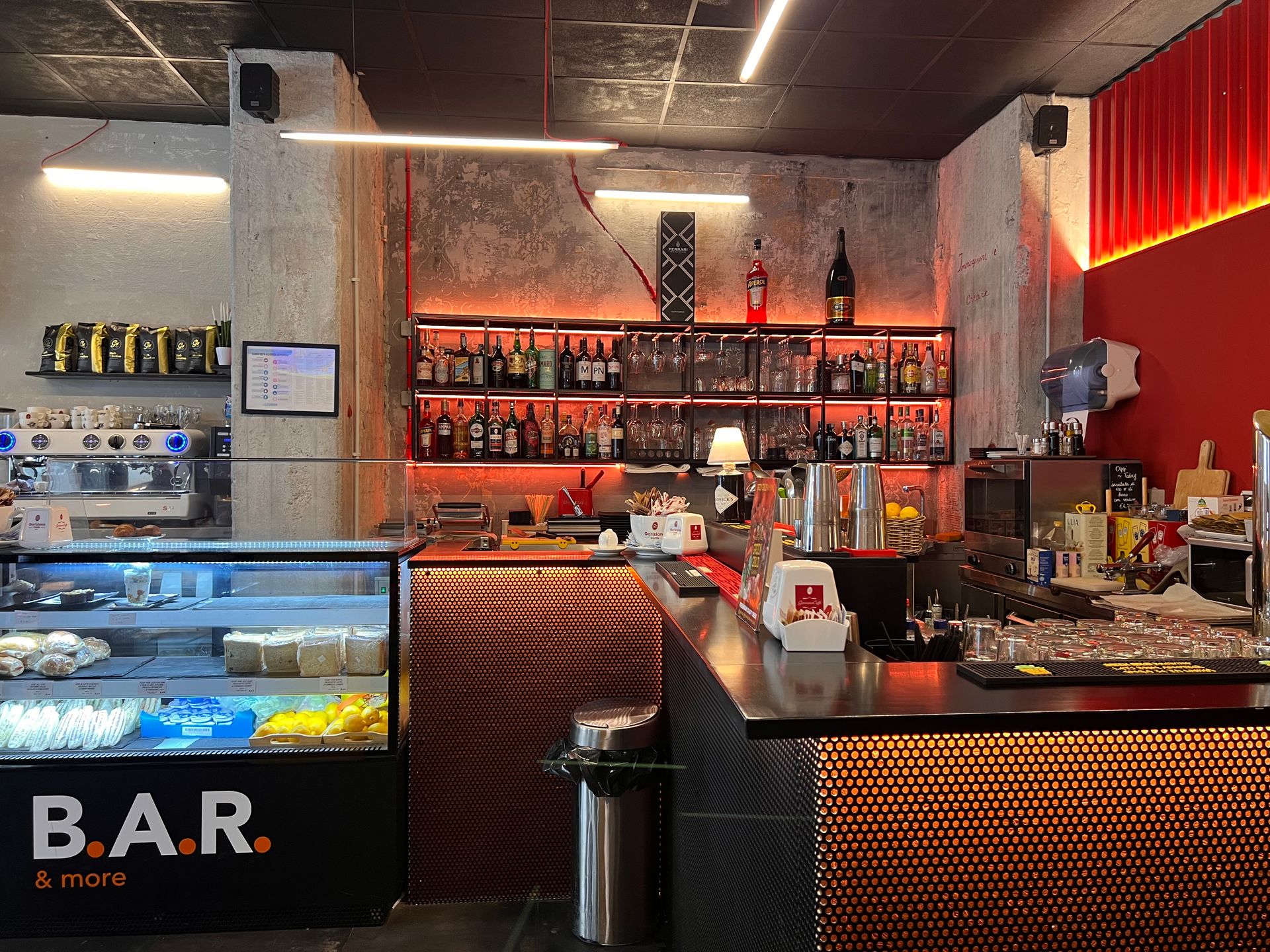 Interno del bar con macchina per l'espresso, bancone, bottiglie di liquori ed esposizione di cibo.