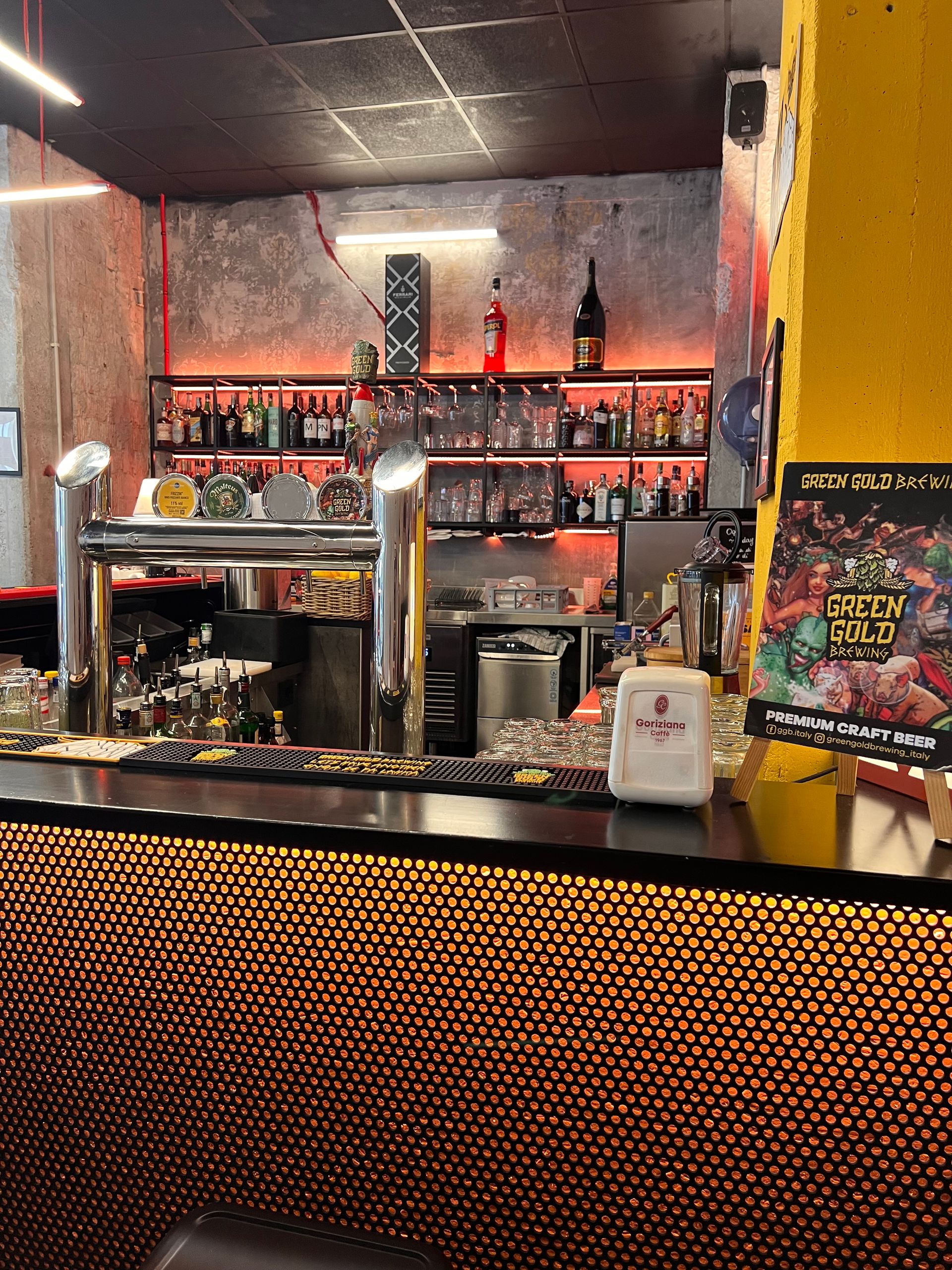 Interno del bar con bottiglie sugli scaffali, rubinetti e facciata strutturata. Illuminazione rossa e gialla.