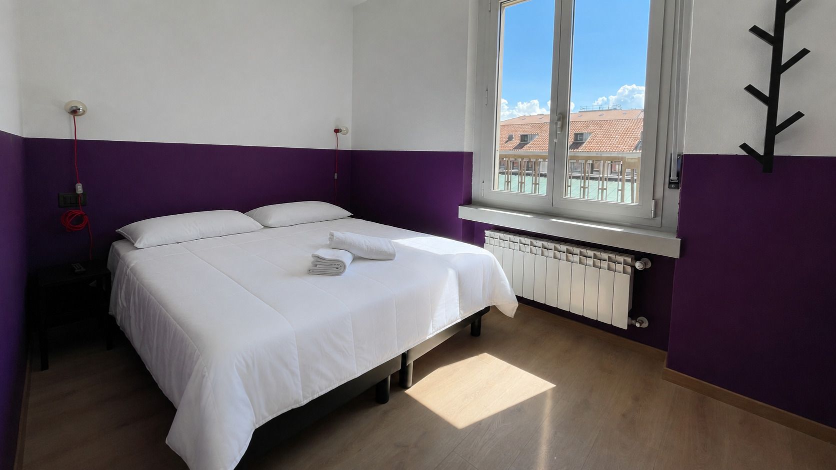 Camera da letto con pareti viola, letto matrimoniale, finestra con vista, termosifone e attaccapanni.