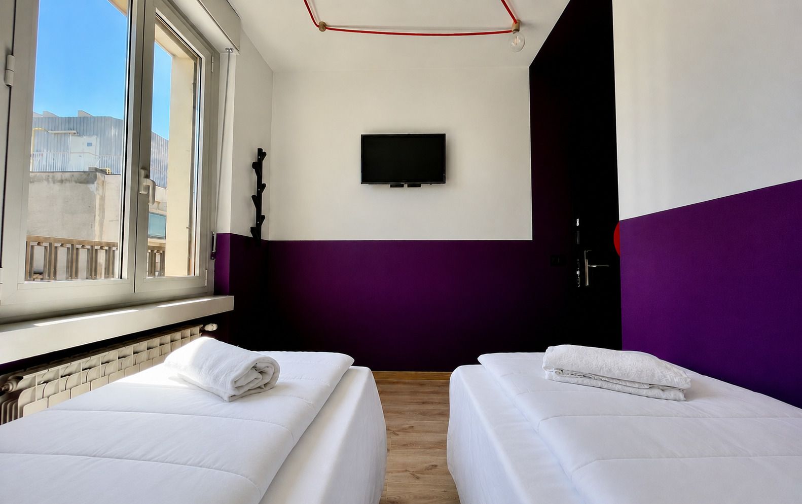 Due letti singoli in una stanza con una parete viola e bianca, una finestra e una TV.