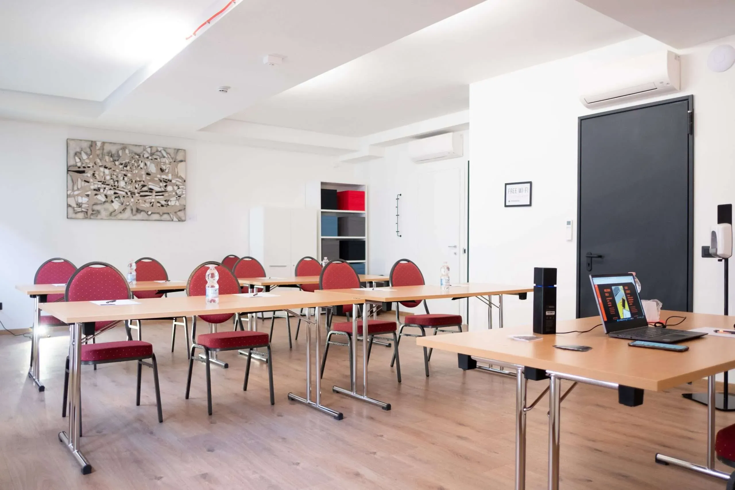 Aula con sedie rosse, tavoli, pareti bianche e una porta nera.