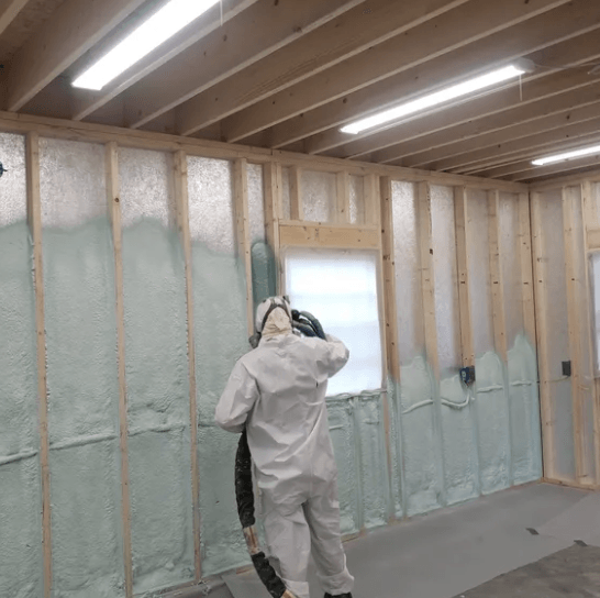 Person in protective suit spraying foam insulation on a wall inside a building.