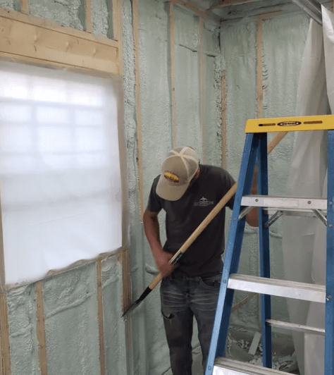 Person using a shovel to clean excess foam from a wall near a window and ladder. Inside a room.