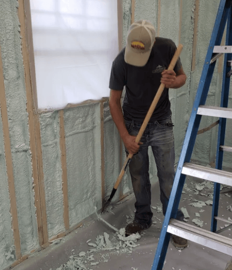 Person scrapes excess insulation from a floor in a room with a window, ladder, and partially insulated walls.