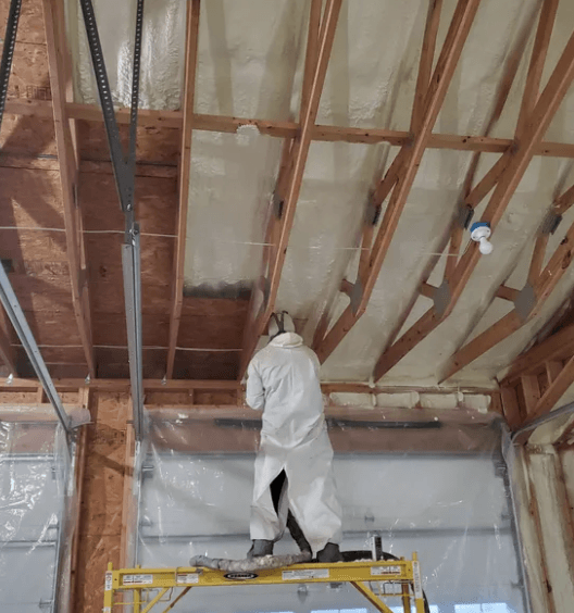 Person in white protective suit spraying insulation onto a ceiling. Scaffolding is below.