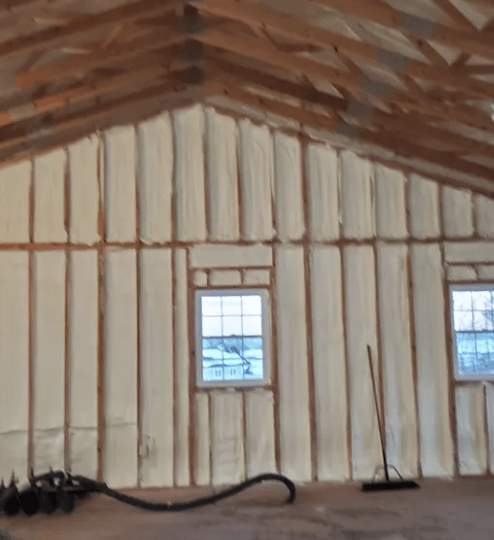 Interior of a building being insulated with spray foam; wood framing, windows, and tools are visible.