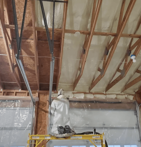 Person in protective suit spraying foam insulation on a ceiling with exposed wooden beams.