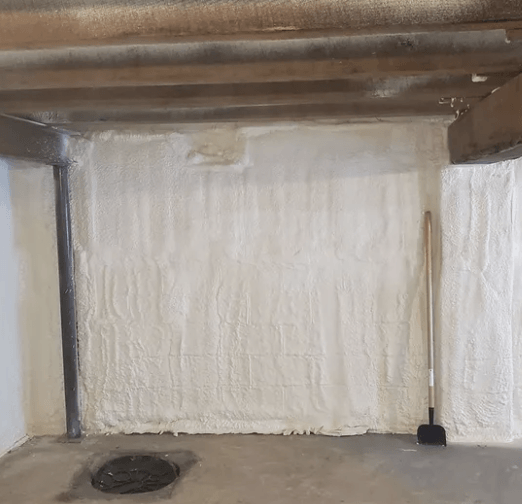 Basement wall insulated with white spray foam, with a shovel leaning against the wall.