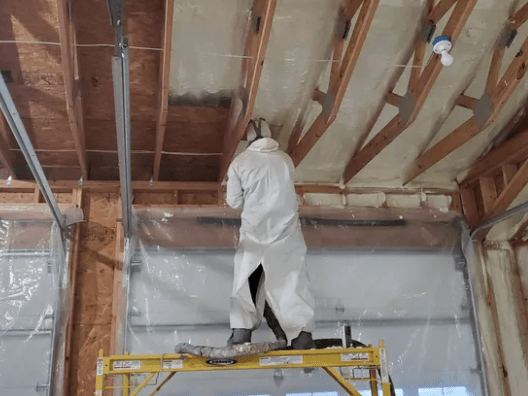 Person in protective suit spraying foam insulation on a wall inside a building.