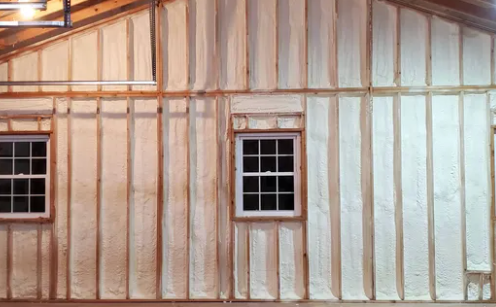 Interior wall with white spray foam insulation between wooden studs, two windows.