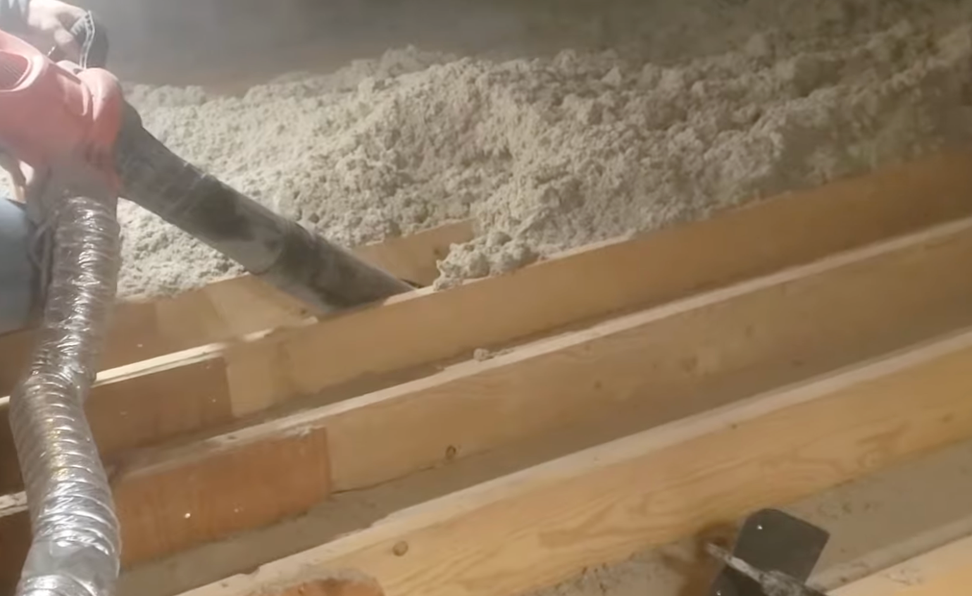 Person using a hose to fill space between wooden beams with insulation.