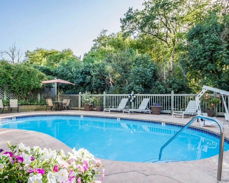 Swimming pool surrounded by a white fence, lounge chairs, and lush greenery on a sunny day.