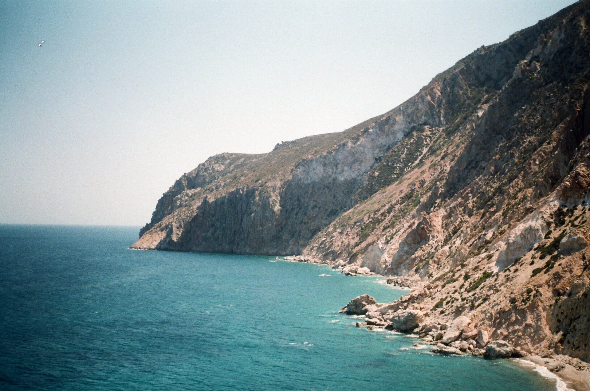 Ocean meets steep, rocky cliffside under a clear, blue sky.