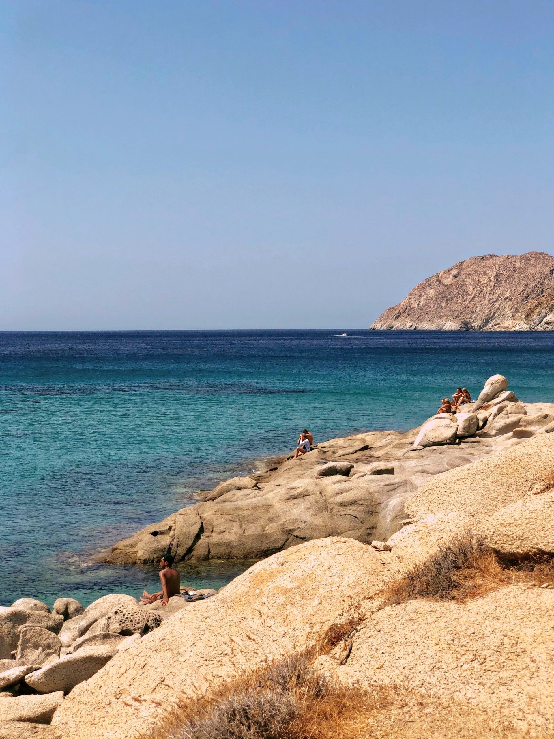 Rocky shoreline with turquoise water, blue sky, and distant island. People are seen relaxing.