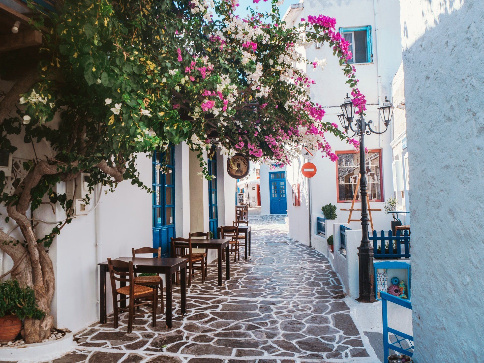 Narrow cobblestone street lined with white buildings, blue doors, and tables under flowering trees.