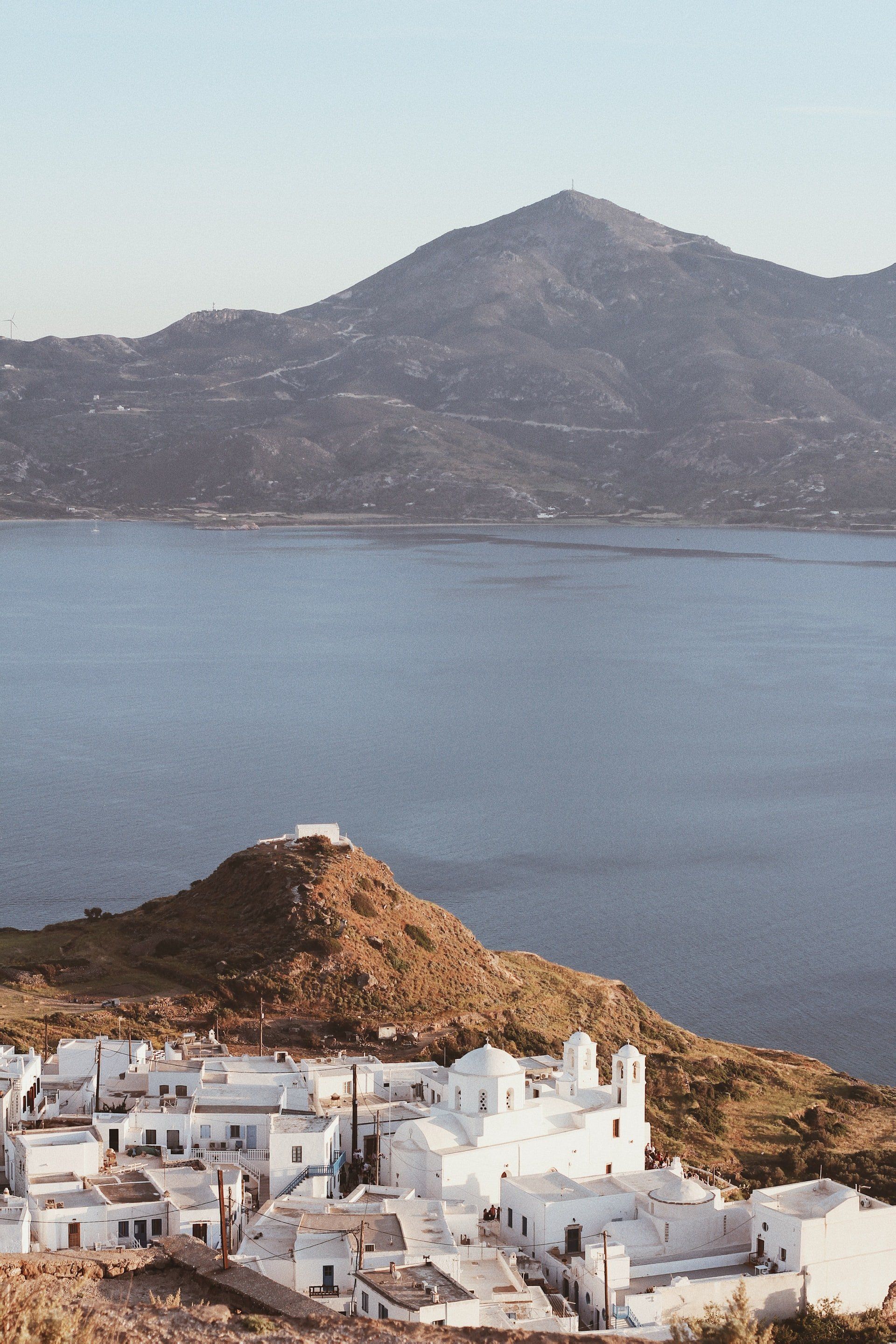 White buildings on a hill, overlooking a blue sea with a large mountain in the background.