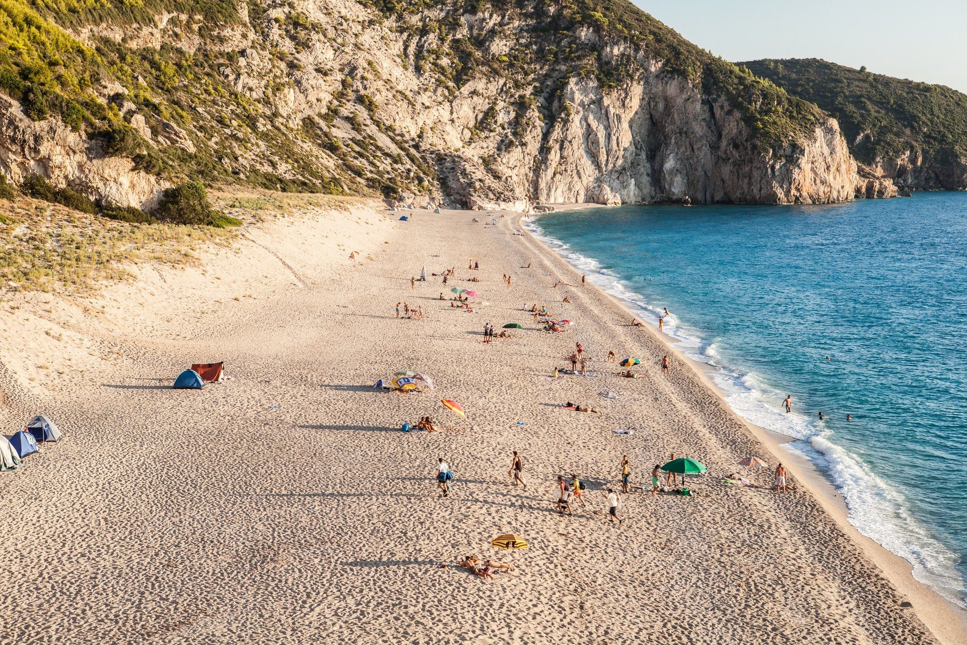 Sandy beach with people sunbathing and swimming; cliffs in the background; blue water.