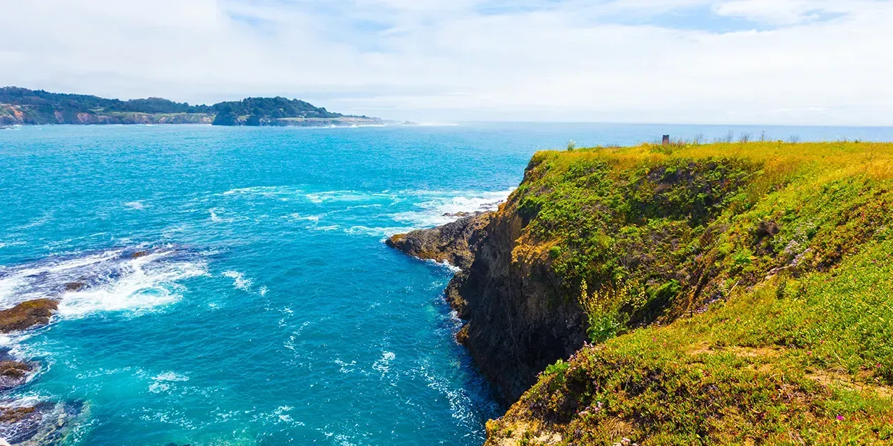 Ocean waves crashing against a rocky coastline, with a grassy cliff in the foreground and a distant town.
