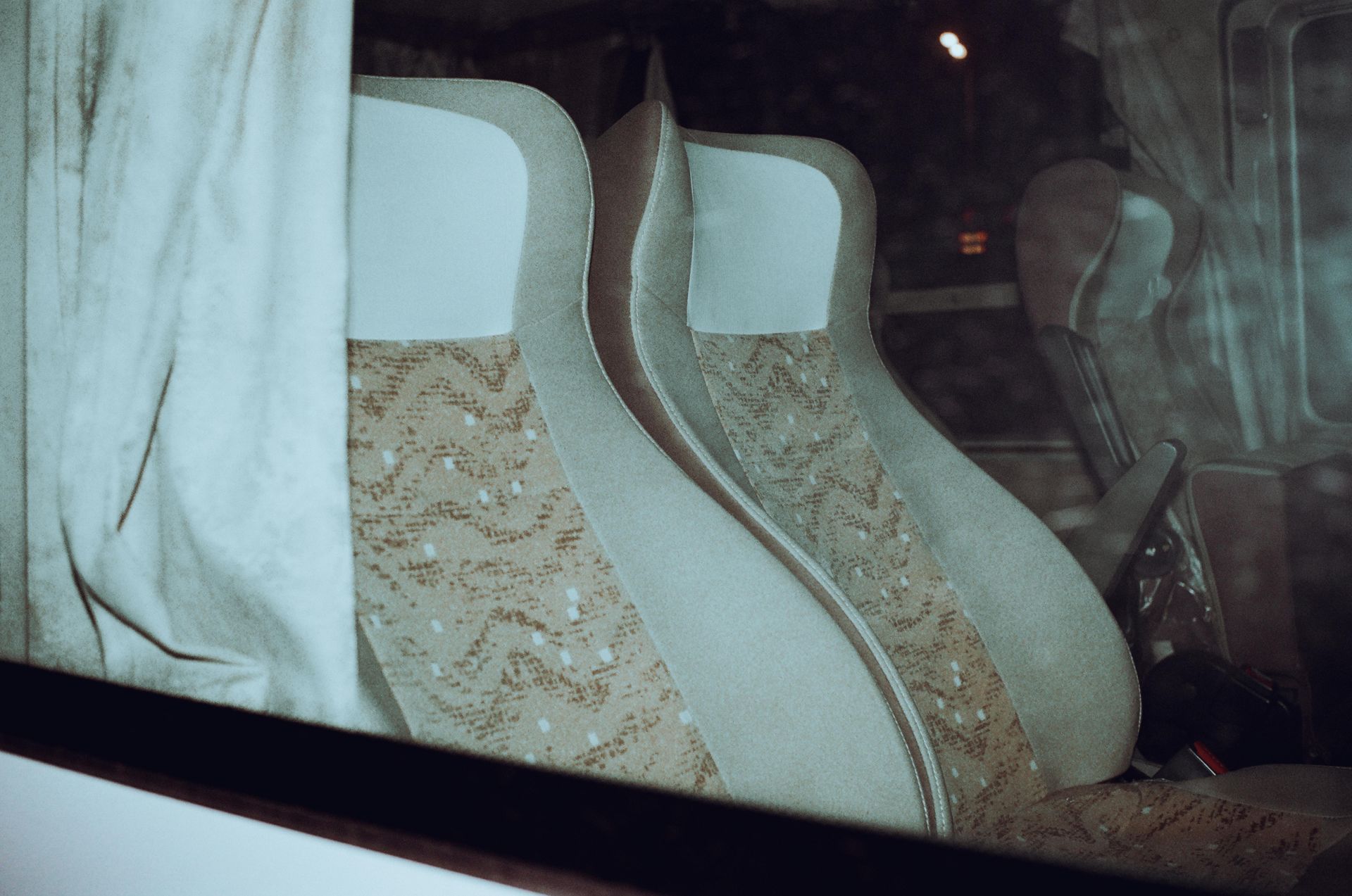 Two empty bus seats, light-colored with patterned fabric, viewed through a window.