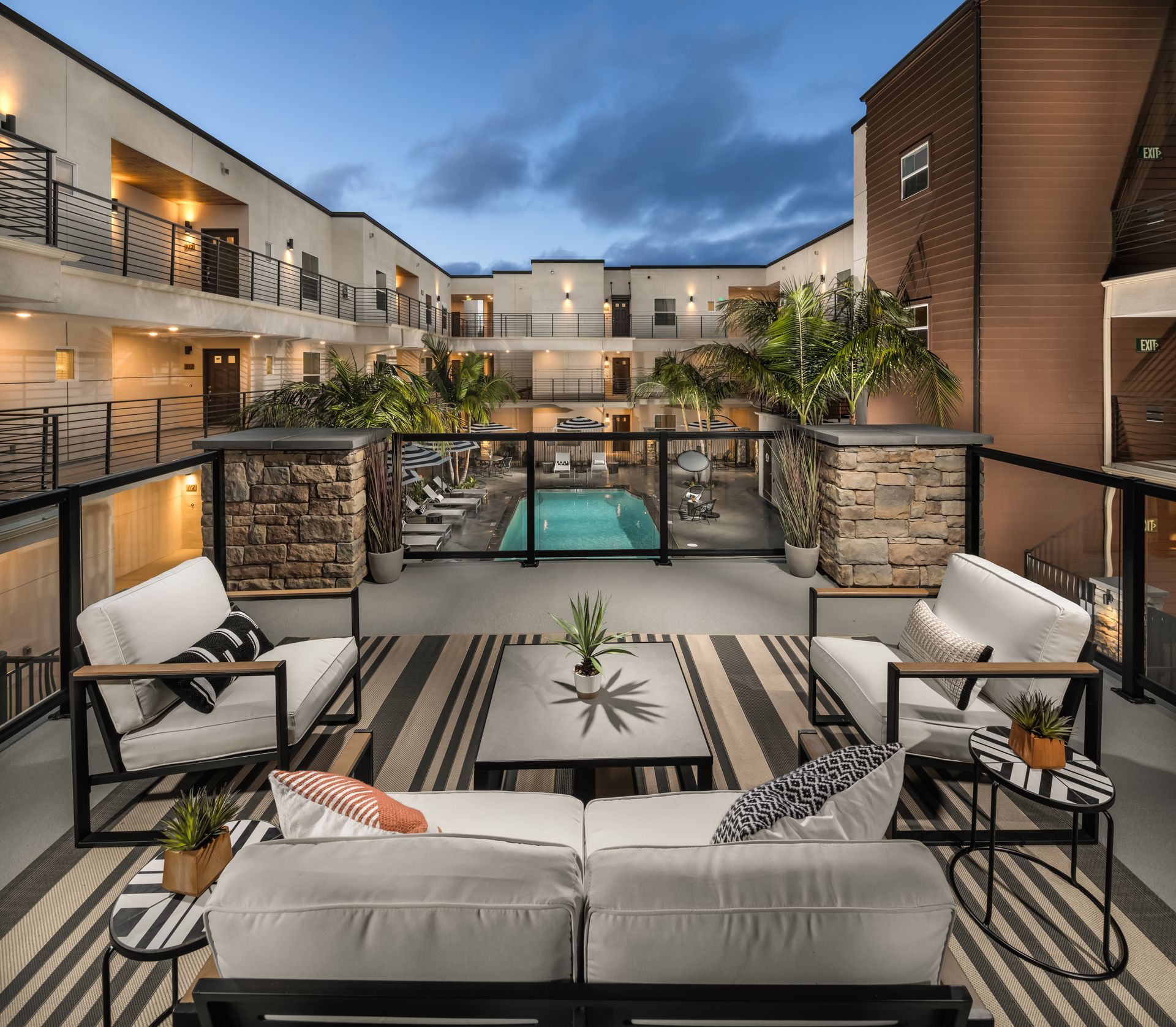 A balcony with a couch , chairs , and a table with a pool in the background.