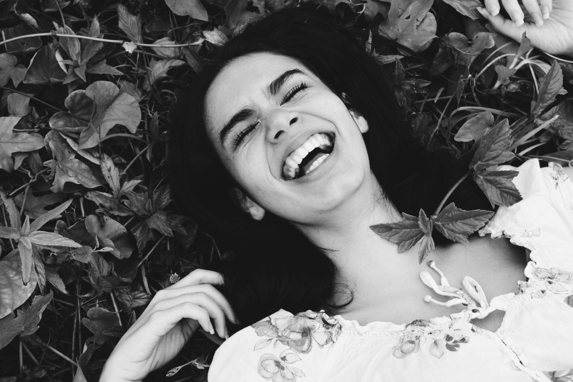 Une personne souriante aux cheveux noirs est allongée dans un tapis de végétation, riant les yeux fermés.