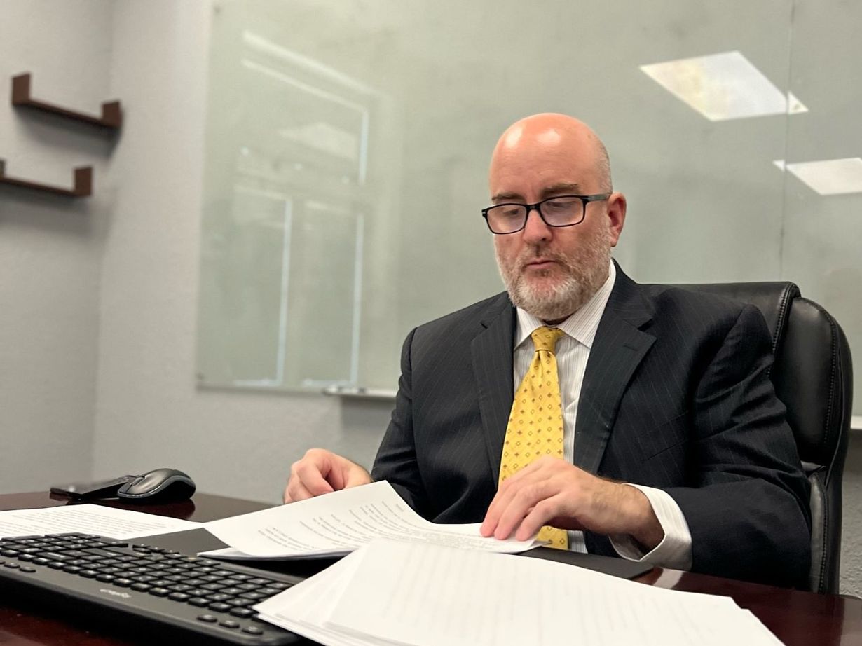 Man in a dark suit and yellow tie reviewing papers at a desk in an office