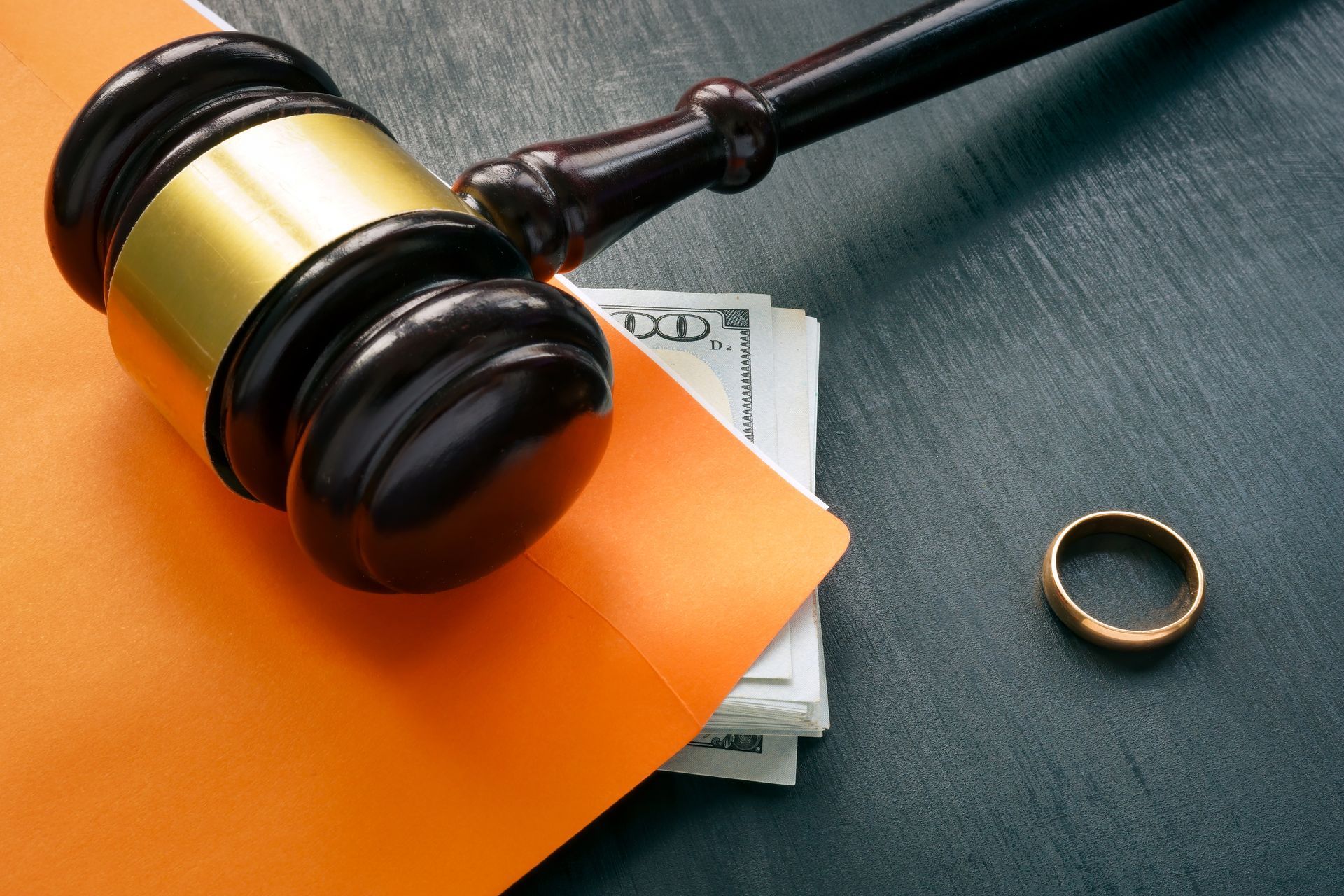 Gavel on an orange envelope beside cash and a wedding ring on a dark surface