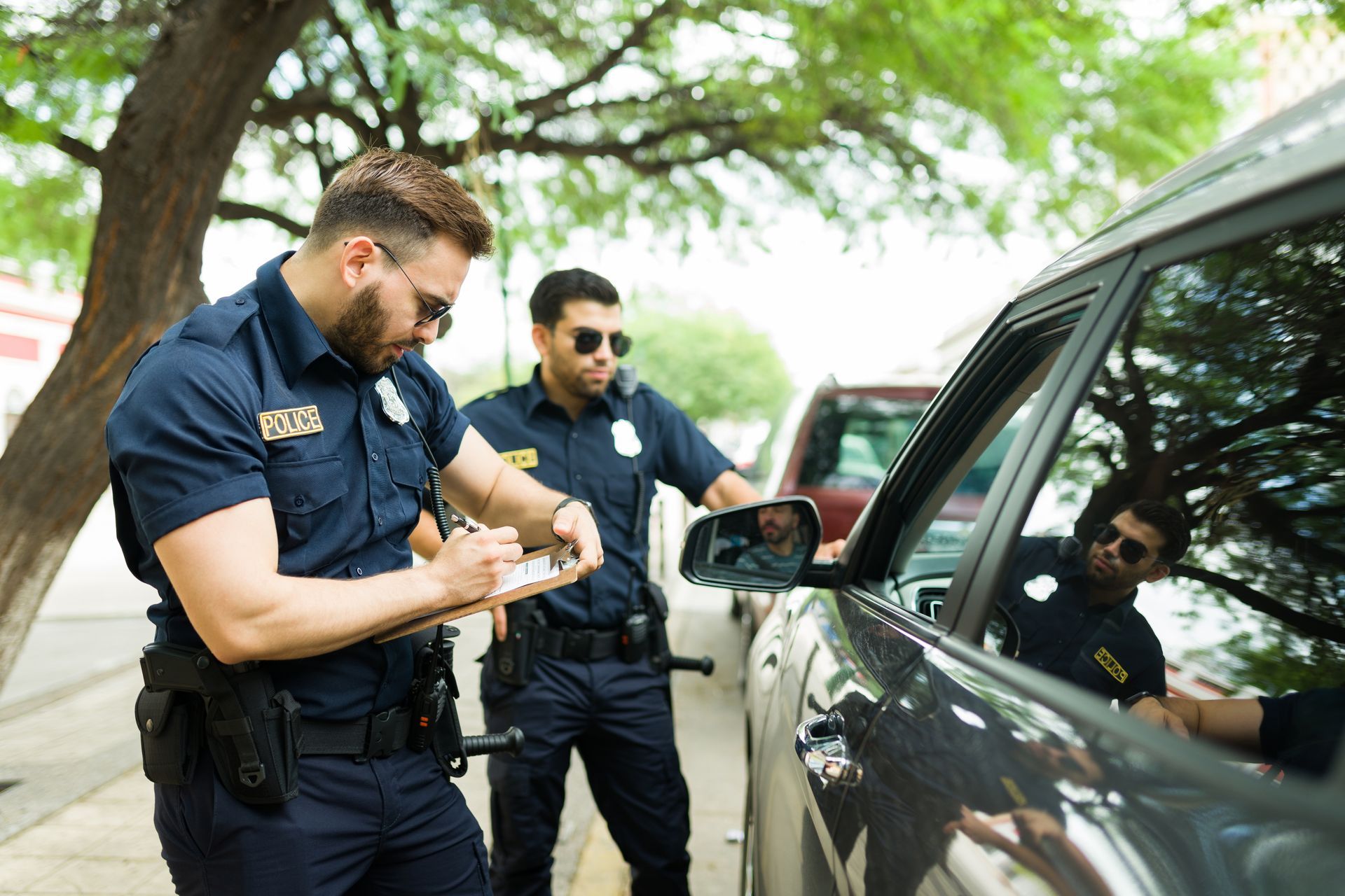 Two police officers speaking with a driver beside a car on a tree-lined street.