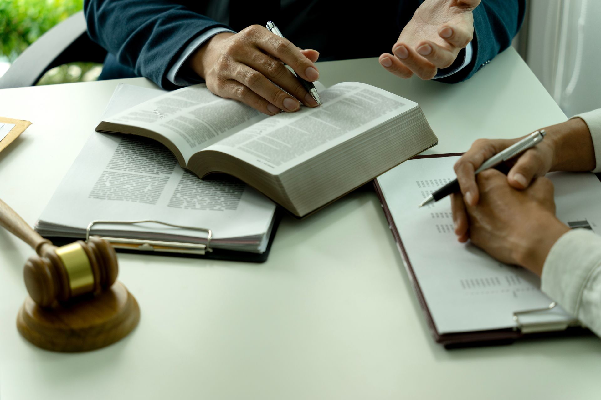 Two people take notes at a table with an open law book and a gavel in the foreground
