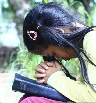 Girl with clasped hands bowed over a book outdoors, wearing a yellow sweater and hair clip