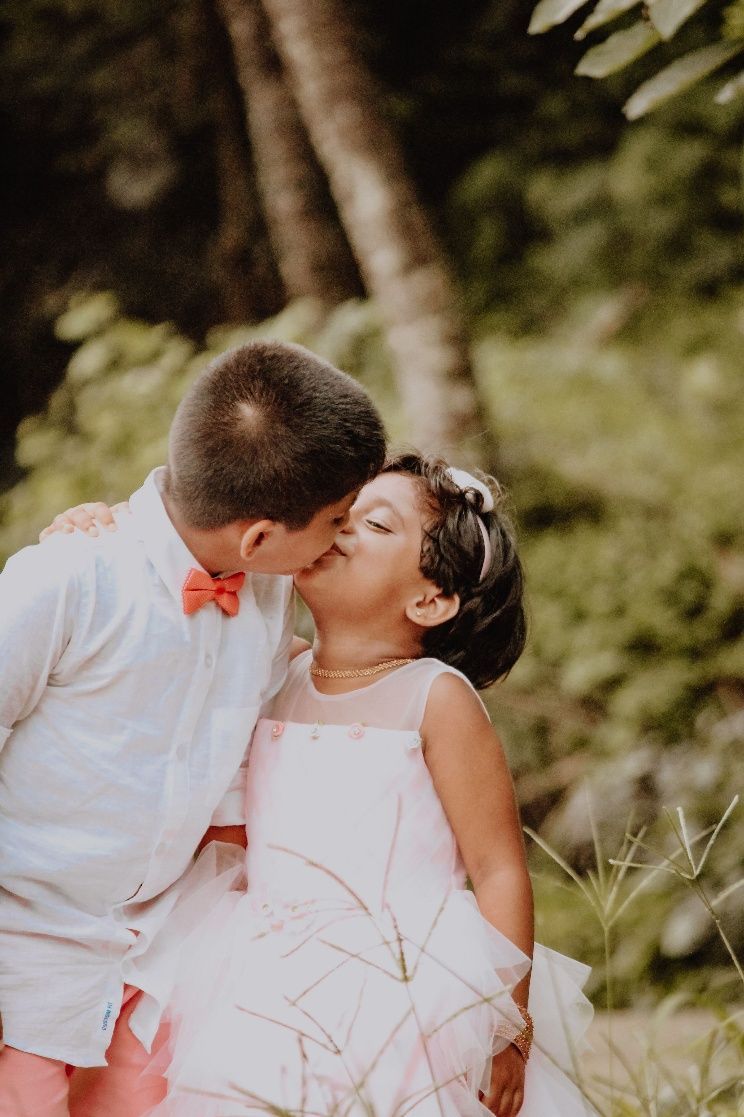 A couple kissing outdoors, with the woman in a pink dress and the man in a white shirt and orange bow tie.