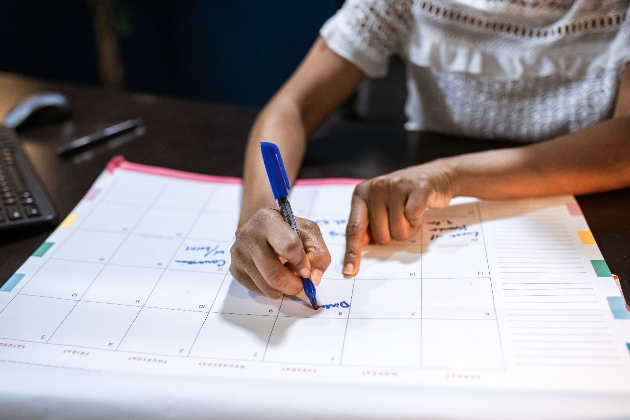 Hands writing on a paper calendar with a blue pen beside a keyboard and laptop